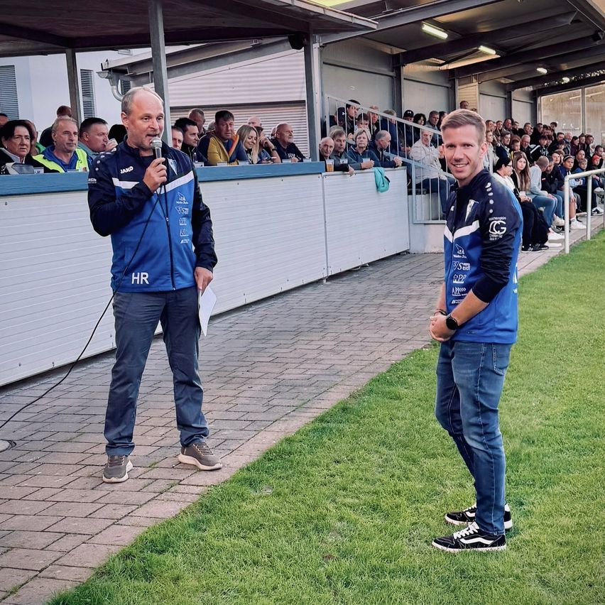 Zwei Männer stehen auf einem Sportfeld, mit einer Menschenmenge im Hintergrund. Ein Mann hält ein Mikrofon und spricht, während der andere neben ihm steht.