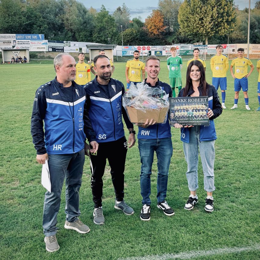 Vier Personen stehen auf einem Fußballfeld, eine Frau hält eine Plakette und eine Geschenkbox. Dahinter stehen eine Gruppe von Fußballspielern in gelben und grünen Uniformen.