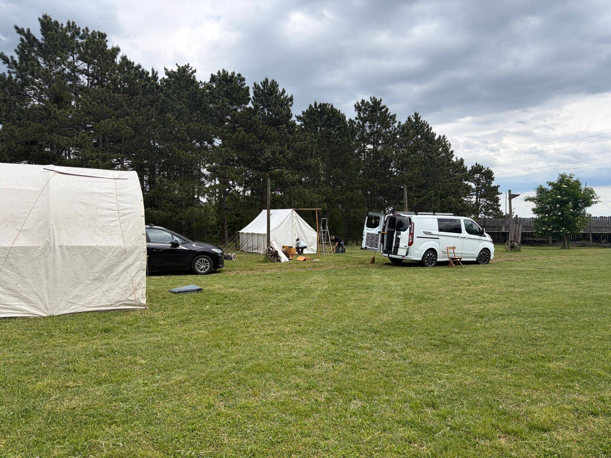 Ein grasbewachsenes Feld mit mehreren Zelten und einem weißen Van mit offenen Türen. Es gibt eine Person, die auf einem Stuhl sitzt, und eine weitere Person, die in der Nähe des Zeltes steht. Im Hintergrund befinden sich mehrere Bäume.