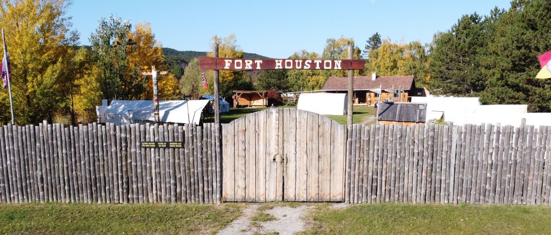 Fort Houston ist eine historische Stätte mit einem Holzzaun und Tor, umgeben von Zelten und Bäumen.