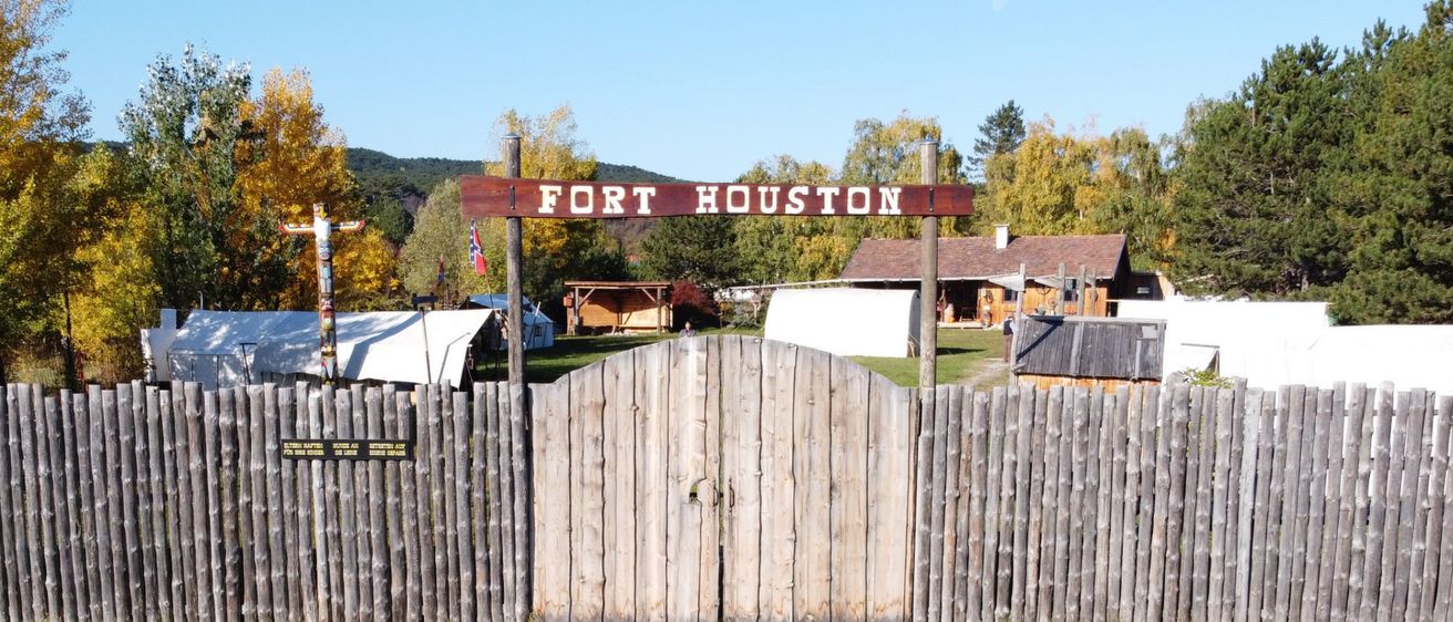 Fort Houston steht auf einem hölzernen Schild über einem offenen Tor. Im Hintergrund sind Zelte und kleine Gebäude zu sehen.