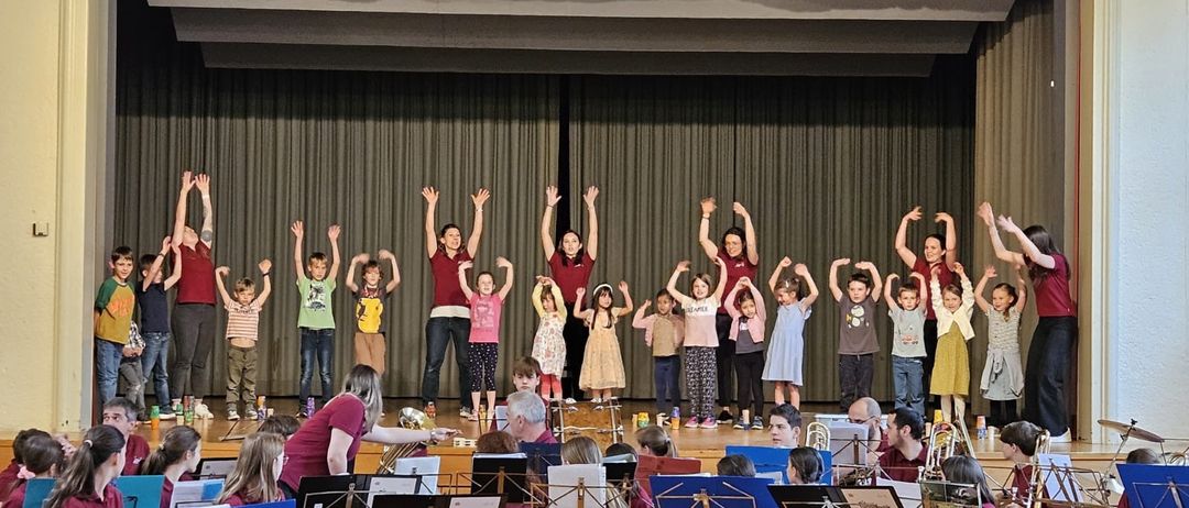 Eine Gruppe von Kindern in passenden roten Shirts tritt auf der Bühne auf, begleitet von Erwachsenen, die Instrumente spielen. Die Kinder heben ihre Hände in die Luft.
