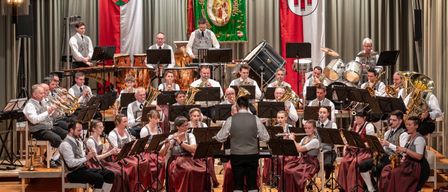 Ein Orchester spielt auf der Bühne mit verschiedenen Instrumenten, darunter Trompeten, Trommeln und Flöten. Die Musiker sind in traditioneller Kleidung gekleidet. Dahinter hängen rote und weiße Flaggen.