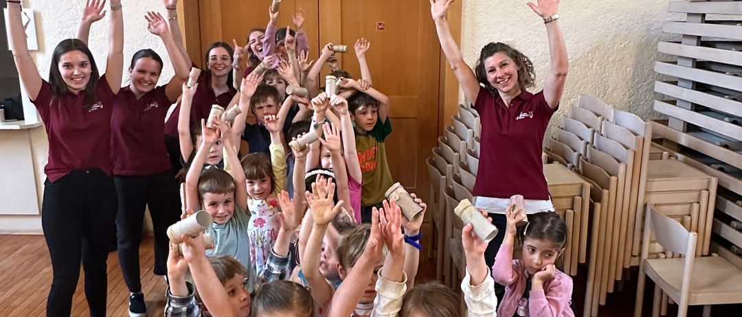 Eine Gruppe von Kindern und Erwachsenen hebt ihre Hände und lächelt in einem Raum mit Holzböden und Stühlen. Einige halten Tassen.