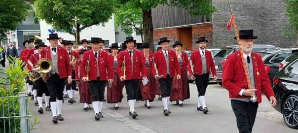 Eine Musikkapelle in roten Uniformen spielt Instrumente, während sie die Straße entlanggeht. Sie wird von einer Flagge begleitet und ist von geparkten Autos und Bäumen umgeben.