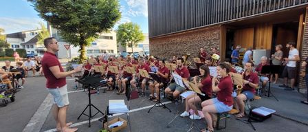 Ein Orchester probet im Freien. Mehrere Musiker sitzen mit Instrumenten, während andere stehen. Ein Dirigent leitet die Probe.