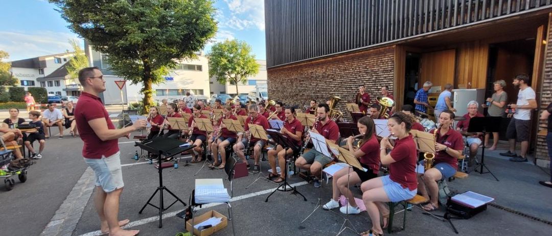 Ein Orchester probet im Freien. Mehrere Musiker sitzen mit Instrumenten, während andere stehen. Ein Dirigent leitet die Probe.