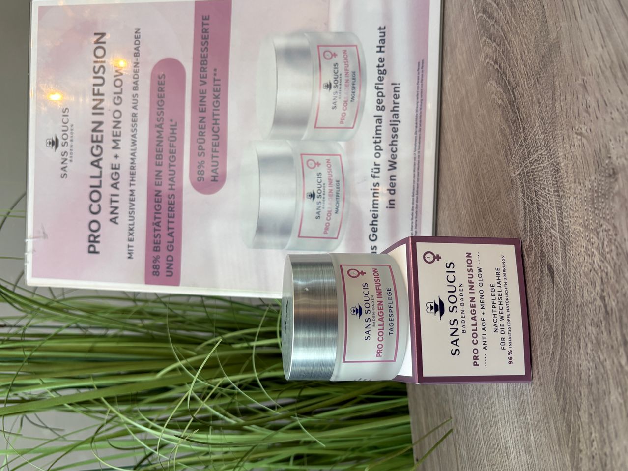 Ein Nahaufnahme einer Kosmetikproduktbox mit silbernem Deckel, die die Marke 'Sans Soucis' und eine Beschreibung von 'Pro Collagen Infusion' zeigt. Die Box steht auf einer Holzoberfläche mit einem Plakat im Hintergrund.