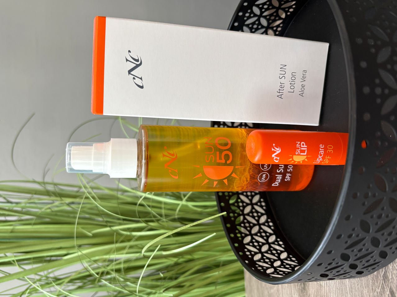 Zwei Flaschen Sonnencreme, eine mit der Aufschrift 'After Sun' und die andere 'Dual Sun SPF 50', liegen auf einem schwarzen Tablett mit einer weißen Schachtel, die 'After SUN Lotion Aloe Vera' beschriftet ist, dahinter.