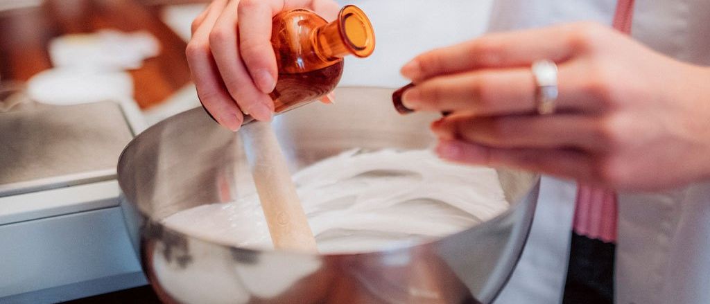 Eine Person fügt eine Flüssigkeit aus einer kleinen Flasche in eine Rührschüssel hinzu, während sie mit einem Holzlöffel rührt. Sie trägt einen Laborkittel und einen Ring.