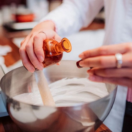 Eine Person in einem Laborkittel fügt mit einem Holzlöffel eine Zutat in eine Schüssel hinzu.