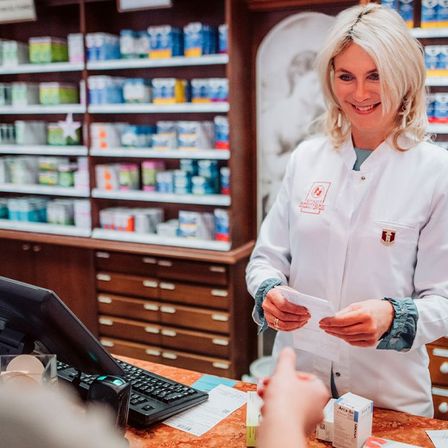 Eine Apothekerin in einem Laborkittel steht an einer Apothekentheke, lächelt und hält ein Stück Papier in der Hand, während sie mit einem Kunden interagiert. Hinter ihr sind Regale mit verschiedenen Medikamenten bestückt.