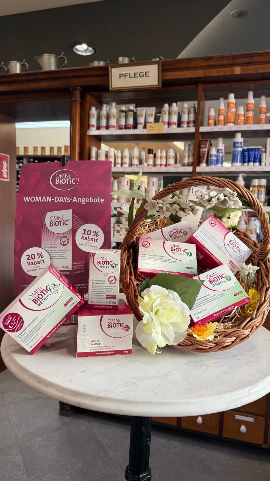 A basket with Omni biotic products and flowers. There are boxes of Omni Biotic Stress, Omni Biotic Meta, and Omni Biotic Skin. The products are displayed in a shop with shelves of beauty products in the background.