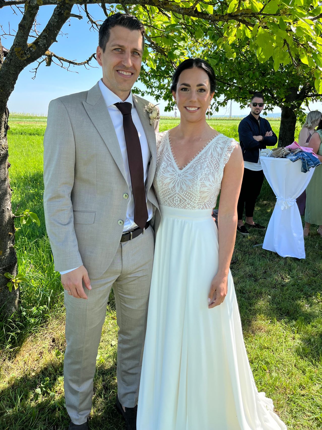 Ein lächelndes Paar posiert für ein Foto auf einem Feld. Der Mann trägt einen Anzug und eine Krawatte, die Frau ein weißes Kleid. Im Hintergrund stehen zwei Personen in der Nähe eines Tisches mit gefalteter Kleidung.