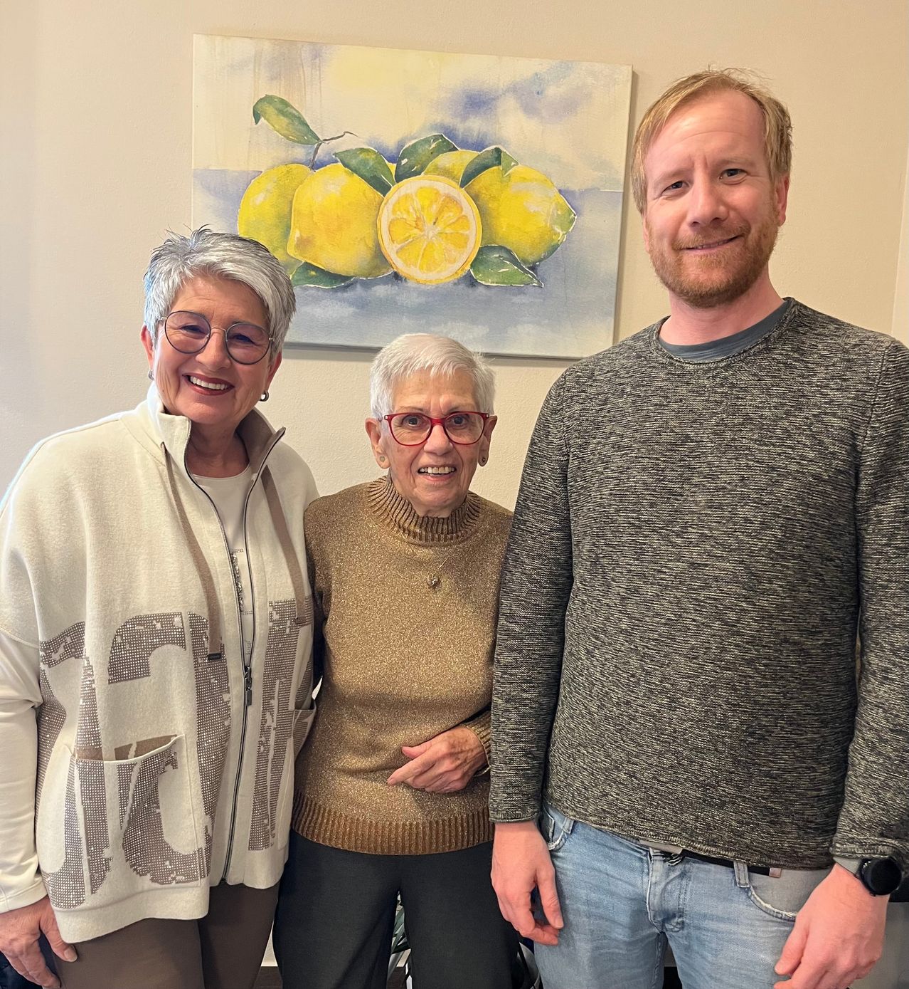 Drei Menschen stehen vor einem Gemälde. Die Frau links trägt eine Brille und eine weiße Jacke mit Buchstaben. Die Frau in der Mitte hat weißes Haar und trägt einen braunen Pullover. Der Mann rechts hat einen Bart und trägt ein graues Langarmshirt.