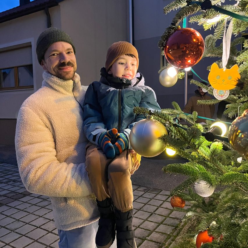 Ein Mann und ein kleiner Junge stehen vor einem geschmückten Weihnachtsbaum und lächeln. Der Mann hält den Jungen auf dem Schoß, und beide sind warm gekleidet.