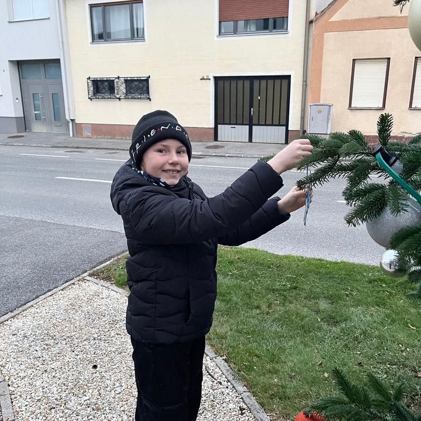 Ein junges Kind in einer Winterjacke und einer Mütze schmückt draußen einen Weihnachtsbaum. Das Kind hält eine Dekoration und platziert sie auf dem Baum. Im Hintergrund befinden sich Gebäude und eine Straße.