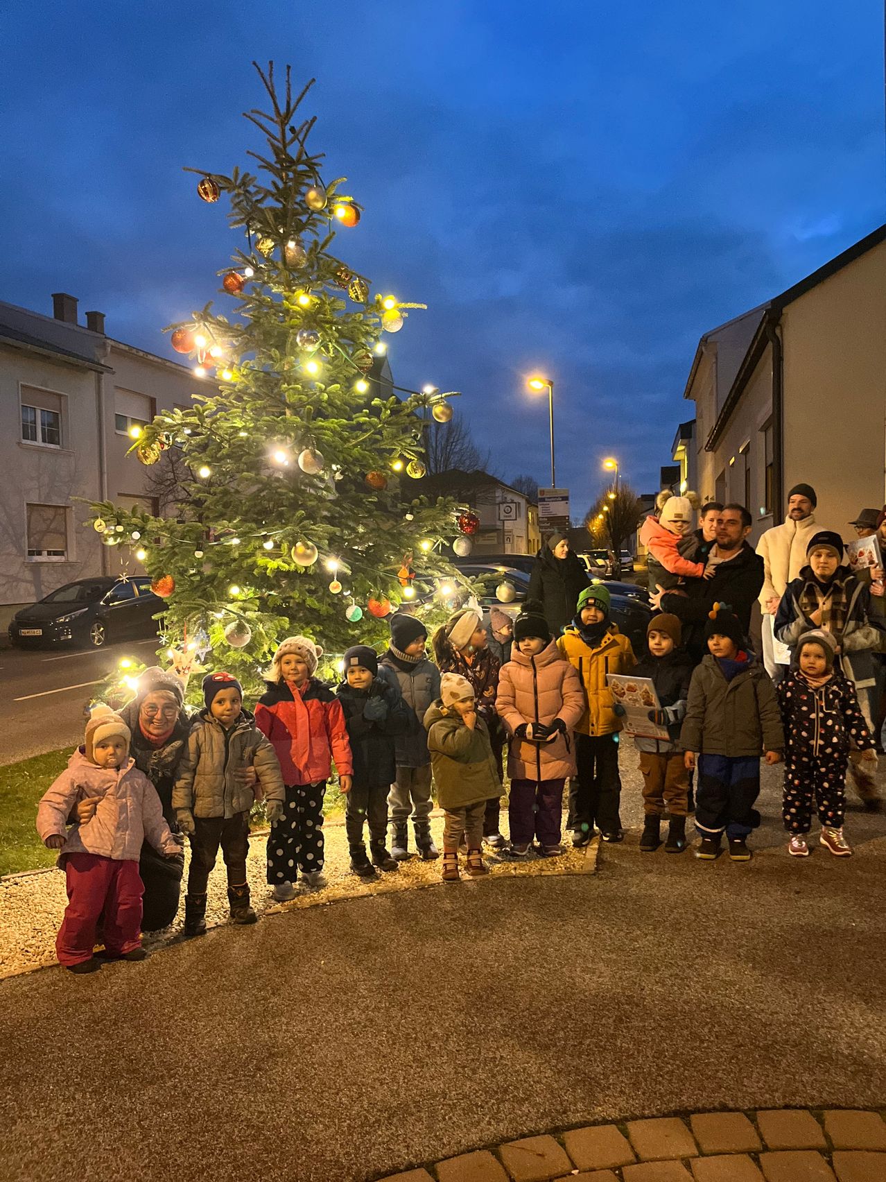 Eine Gruppe von Kindern und Erwachsenen steht vor einem beleuchteten Weihnachtsbaum, der mit bunten Ornamenten geschmückt ist. Sie tragen Winterkleidung. Hinter dem Baum befindet sich ein Gebäude, in der Nähe geparkte Autos. Der Himmel ist bewölkt.