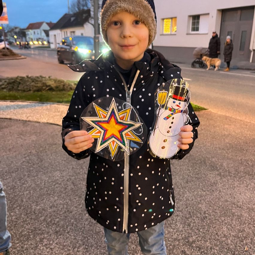 Ein Kind steht nachts auf der Straße, hält eine Stern- und Schneemann-Ausschnitt und trägt eine gepunktete Jacke und Mütze. Dahinter geht ein Hund mit seinem Besitzer, und Gebäude mit eingeschalteten Lichtern sind sichtbar.