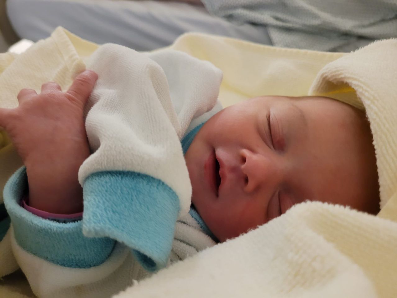 Ein Neugeborenes schläft in einer gelben Decke und trägt ein weiß-blau gestreiftes Outfit. Eine Hand hält sanft die Decke.