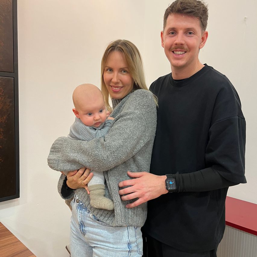 Ein lächelndes Paar hält ein Baby in einem Raum mit weißen Wänden und einem Holzboden. Die Frau trägt einen grauen Pullover und der Mann ein schwarzes Hemd.