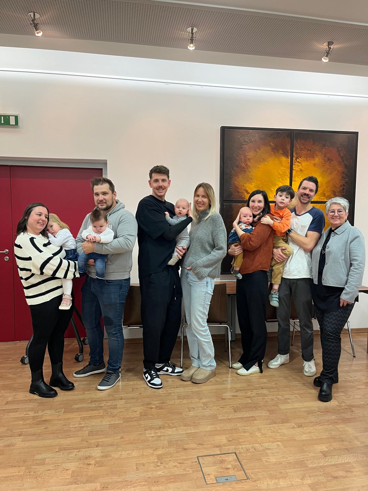 Eine Gruppe von Erwachsenen und Kindern, einschließlich Babys, versammelt sich drinnen und posiert für ein Foto. Einige halten Babys, während andere eng beieinander stehen. Sie befinden sich in einem Raum mit Holzböden, Stühlen und einer Wand mit einem großen Gemälde.