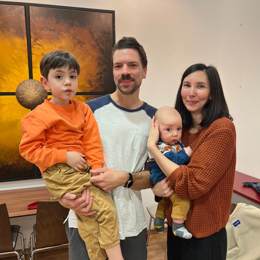 Eine Familie aus drei Personen, bestehend aus einem Mann, einer Frau und zwei Jungen, steht in einem Raum. Der Mann hält einen Jungen in den Armen, während die Frau den anderen Jungen hält. Sie lächeln. Hinter ihnen befindet sich ein Gemälde.