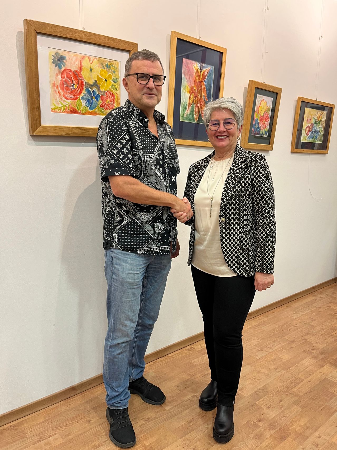 Ein Mann und eine Frau stehen vor einer Wand mit Blumengemälden und schütteln sich die Hände. Der Mann trägt eine Brille und ein gemustertes Hemd, während die Frau eine Brille und einen gemusterten Blazer trägt.