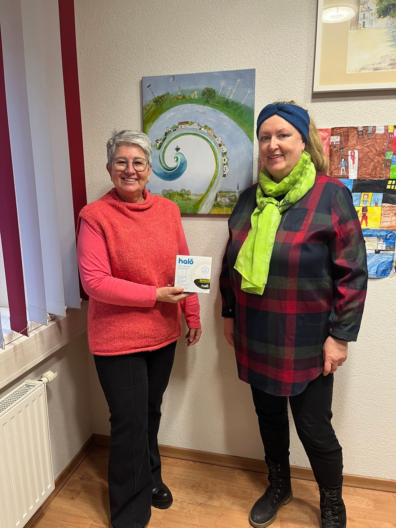 Zwei Frauen stehen in einem Raum mit einem Gemälde dahinter. Eine Frau hält ein Zertifikat und lächelt, während die andere Frau eine blaue Kopfband und eine grüne Krawatte trägt.