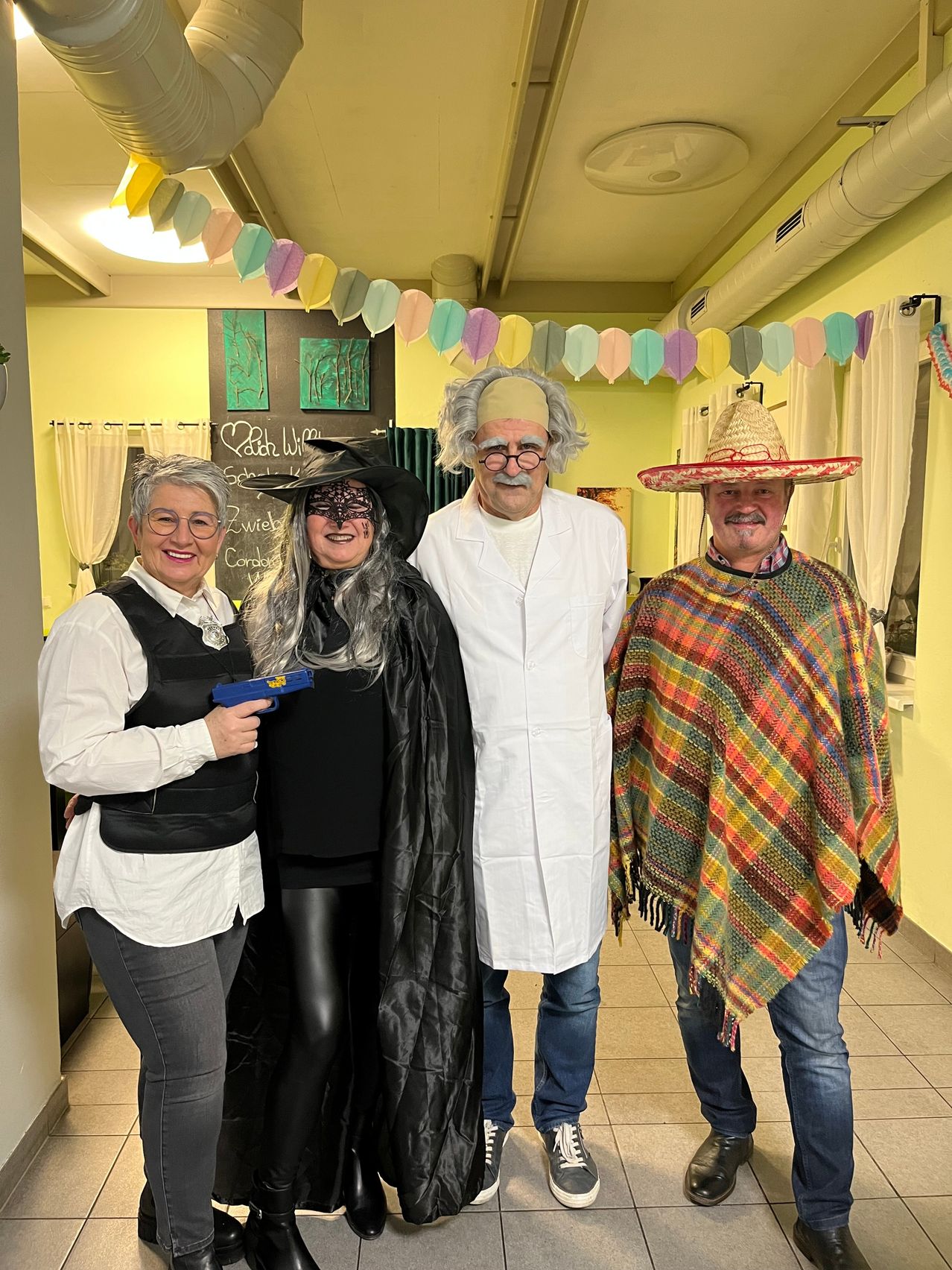 Four people in costumes pose for a photo in a decorated hallway. A woman in a witch costume and a bulletproof vest, a man in a lab coat, and two men in costumes, one wearing a sombrero.