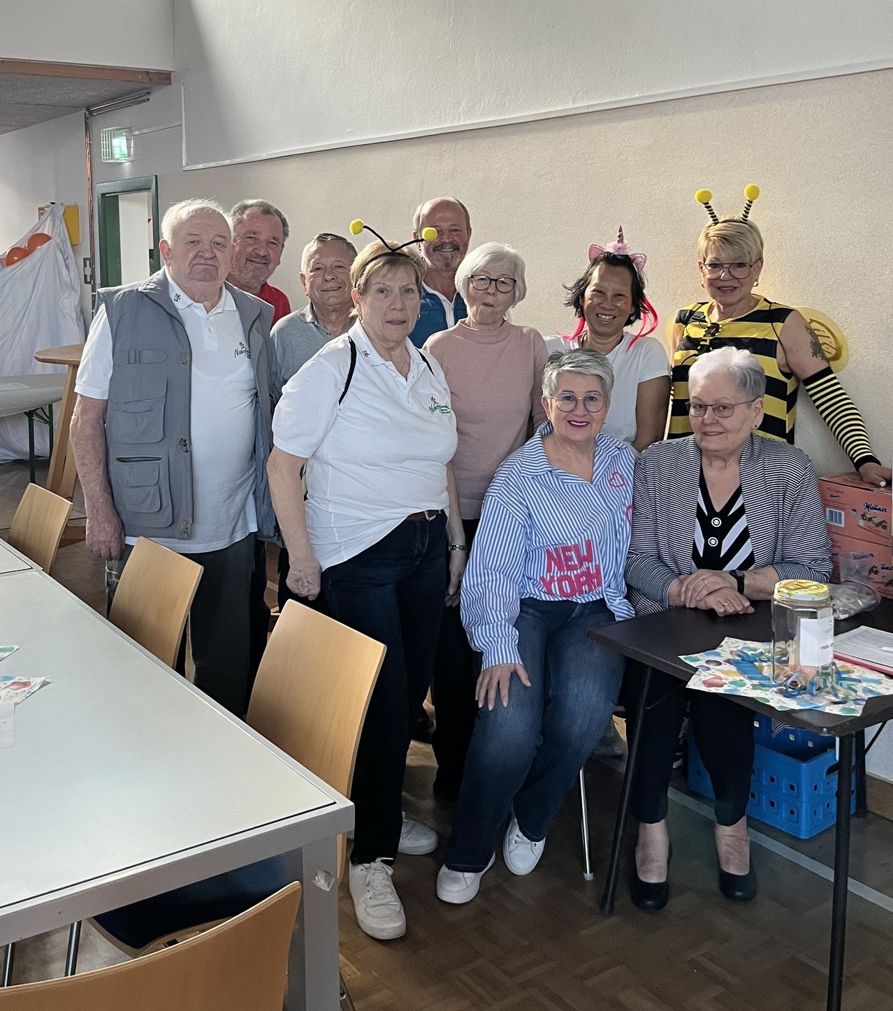 A group of adults, including two women dressed as bees, are posing for a photo in a room with wooden floors and chairs. One woman is wearing a 'New York' shirt. A desk with a jar is in the foreground.