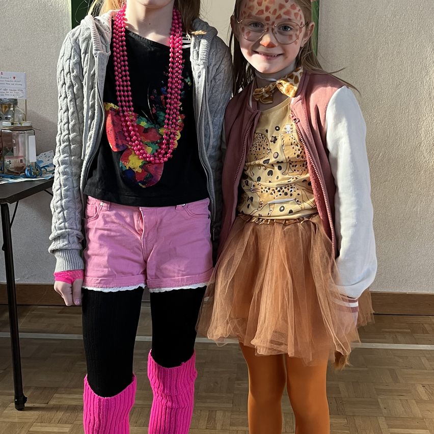 Two girls in costumes pose for a photo. One wears a giraffe face paint, a tutu, and glasses. The other has a pink necklace, boots, and a jacket. Behind them is a table with a jar.