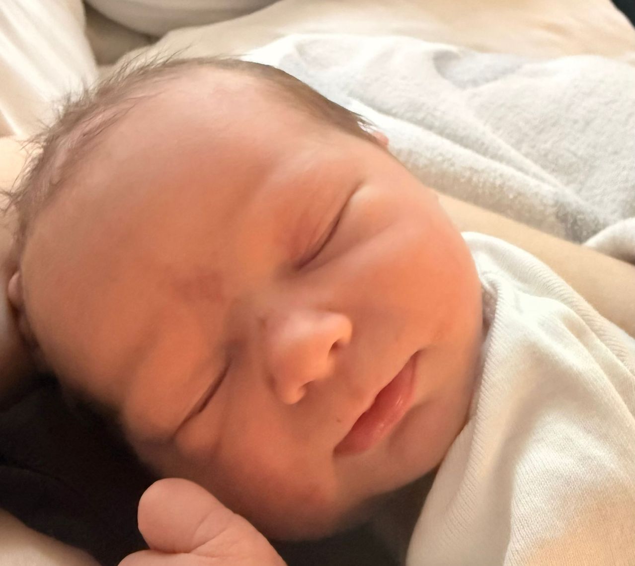 A sleeping newborn baby is being held in someone's arms. The baby is wearing a white outfit and is looking peaceful.