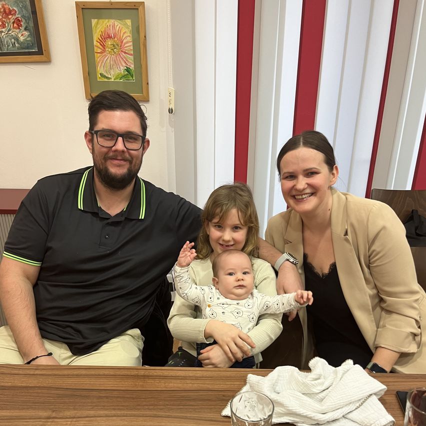 A family of four is seated at a table, smiling for a photograph. The man wears glasses and a black polo shirt, while the woman holds a baby. A young girl stands between them. Behind them, there are two framed paintings on a wall.