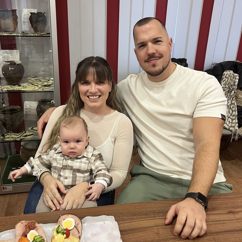 A smiling family sits at a table with food in front of them, a woman holds a baby, and a man stands behind her, all inside a room with a glass cabinet.