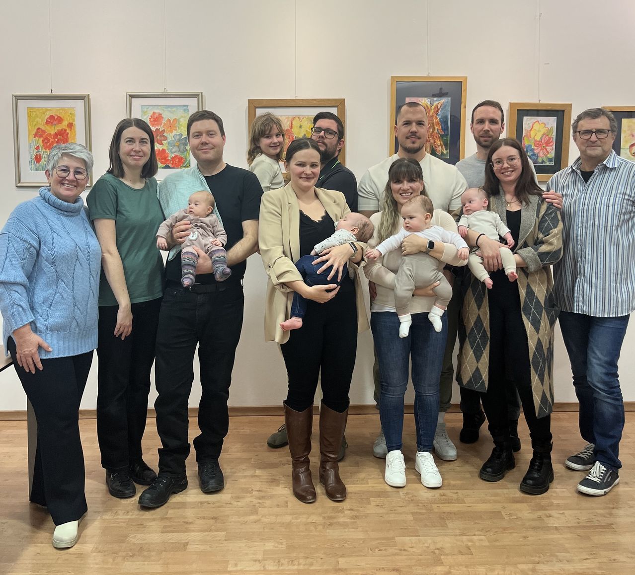 A family gathering in a room with wooden floors and paintings on the wall. Adults and children, including babies, are standing close together smiling for a photo.