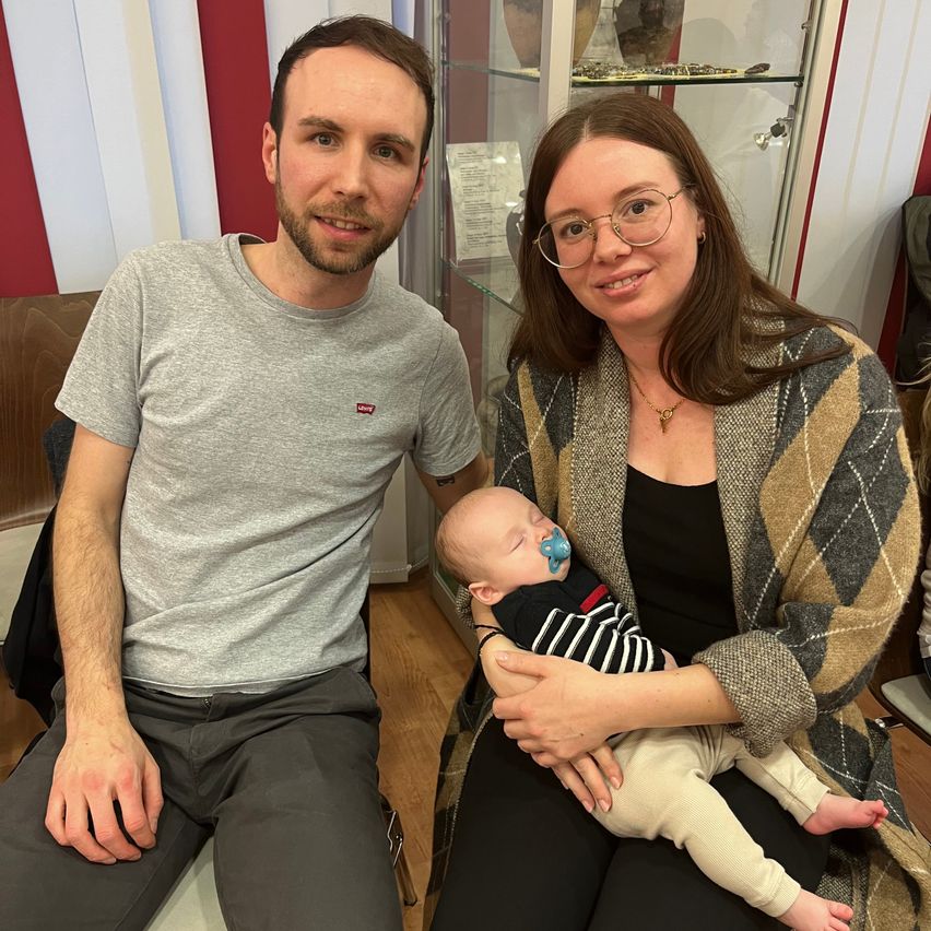 A man and a woman sit on a chair, smiling. The woman holds a sleeping baby with a pacifier in their hand. The baby wears a striped shirt. Behind them is a glass cabinet.