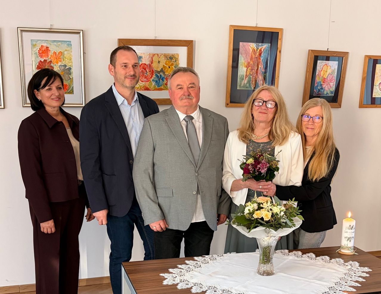 Five individuals, dressed formally, stand in a room with framed paintings on the wall. A woman holds a bouquet of flowers in front of a table with lace.