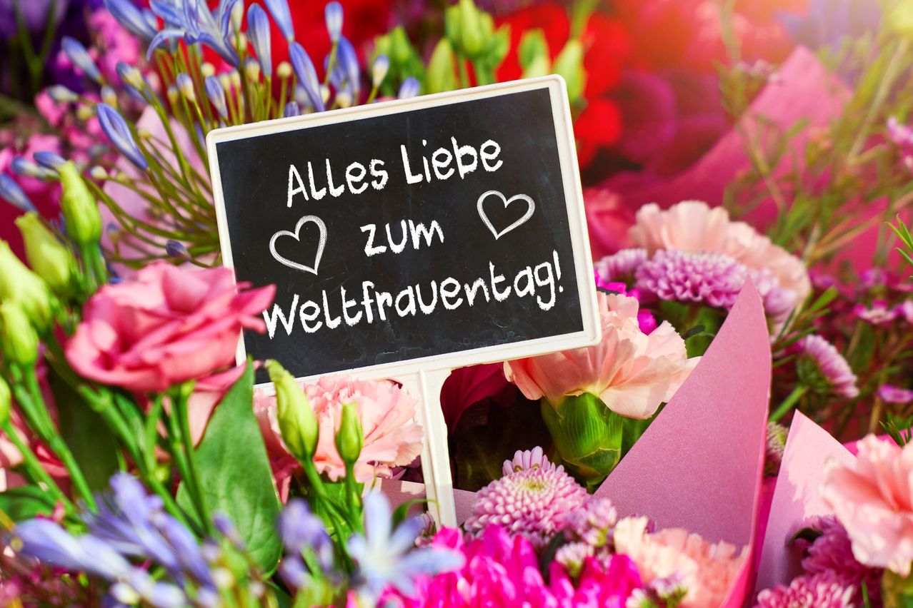 Ein Kreidetafel mit der Aufschrift 'Alles Liebe zum Weltfrauentag!' steht inmitten eines Straußes rosa Blumen und feiert den Internationalen Frauentag.