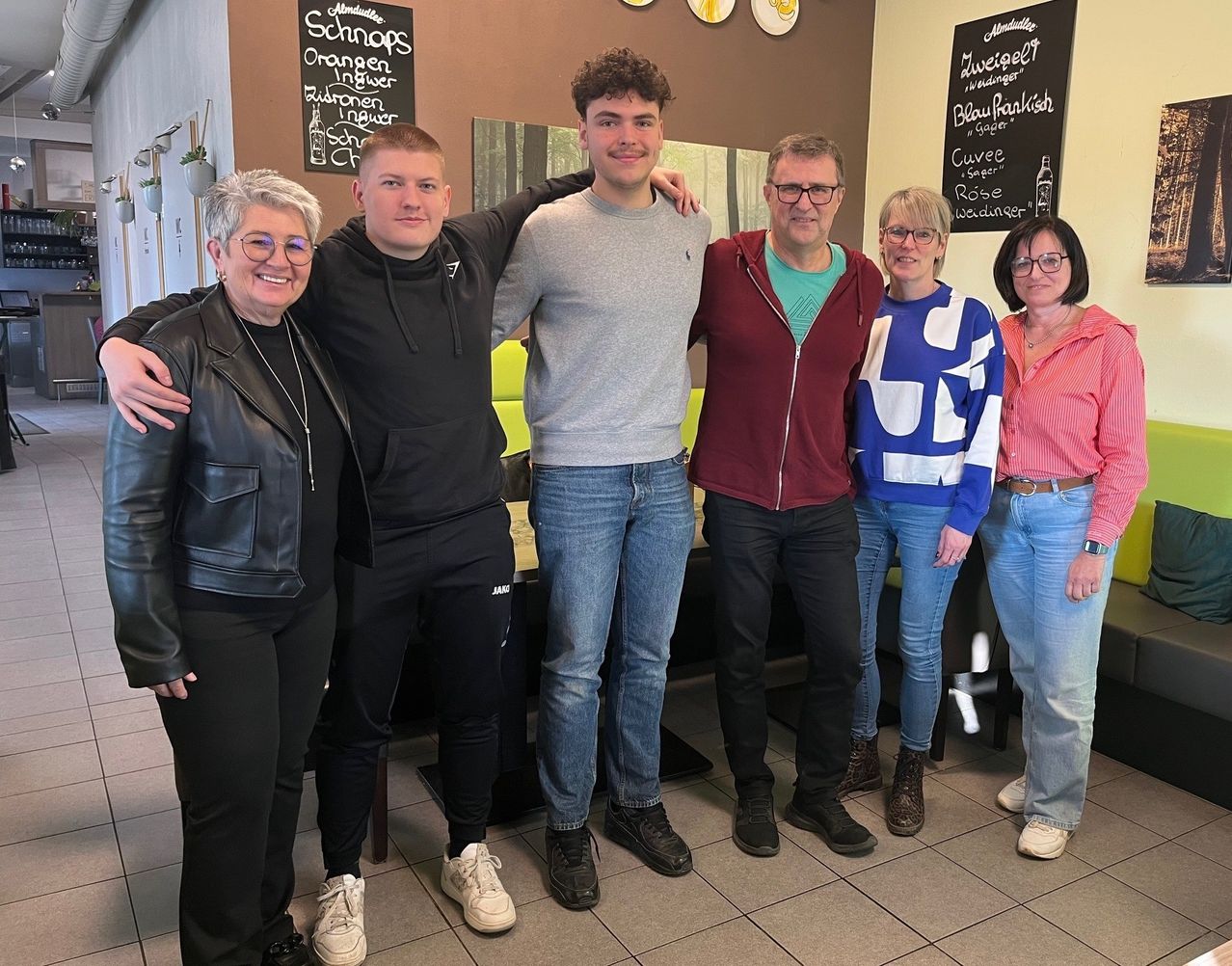 Fünf Personen, darunter zwei Frauen und drei Männer, stehen eng beieinander und posieren für ein Foto. Hinter ihnen befinden sich zwei Tafeln mit Text. Eine von ihnen hat ein Bild einer Flasche.