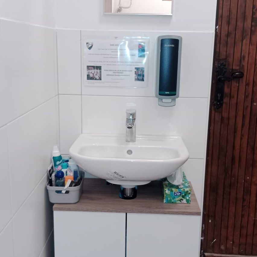 Ein Badezimmer mit einem Waschbecken, Wasserhahn und einer an der Wand montierten Seifenspender. Eine Schachtel Taschentücher und Toilettenartikel stehen auf dem Tisch.