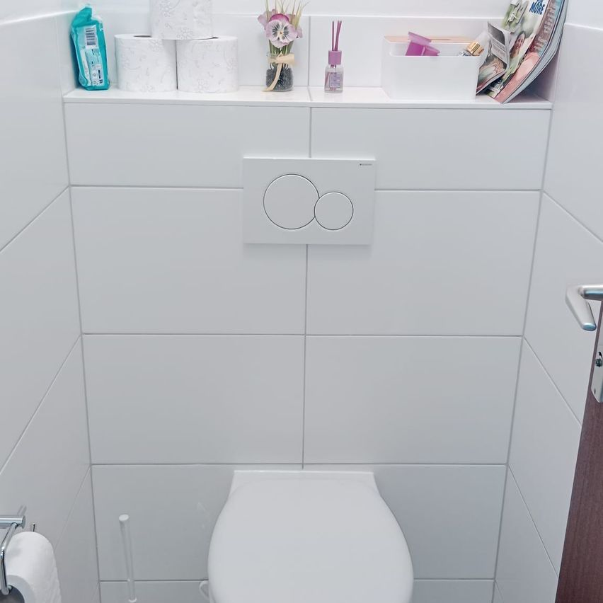 Ein sauberes weißes Badezimmer mit gefliesten Wänden. Eine Toilettenschüssel, ein Regal mit Blumen, Zeitschriften und Toilettenartikeln. Eine Rolle Toilettenpapier an der Wand.