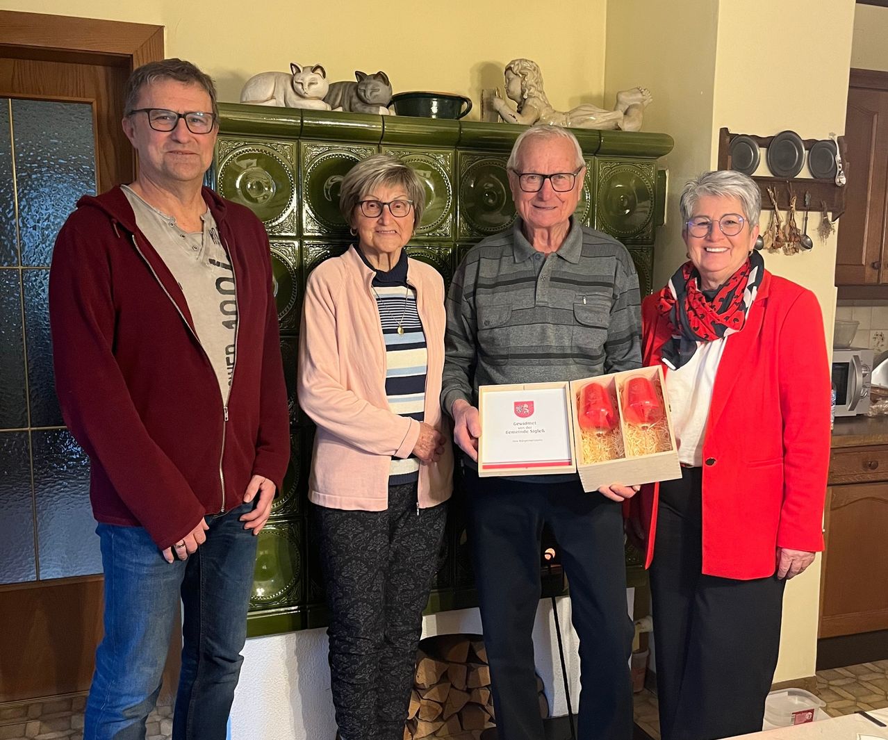 Vier Personen stehen in einem Raum. Ein Mann und eine Frau halten eine Geschenkbox. Eine weitere Frau steht neben ihnen und lächelt. Ein Schrank mit Skulpturen steht hinter ihnen.