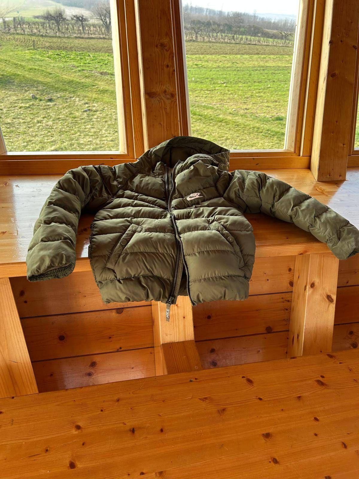 Eine grüne Daunenjacke liegt auf einer Holzbank vor einem Fenster. Die Jacke hat eine Kapuze und einen Reißverschluss. Draußen ist ein grasbewachsenes Feld zu sehen.
