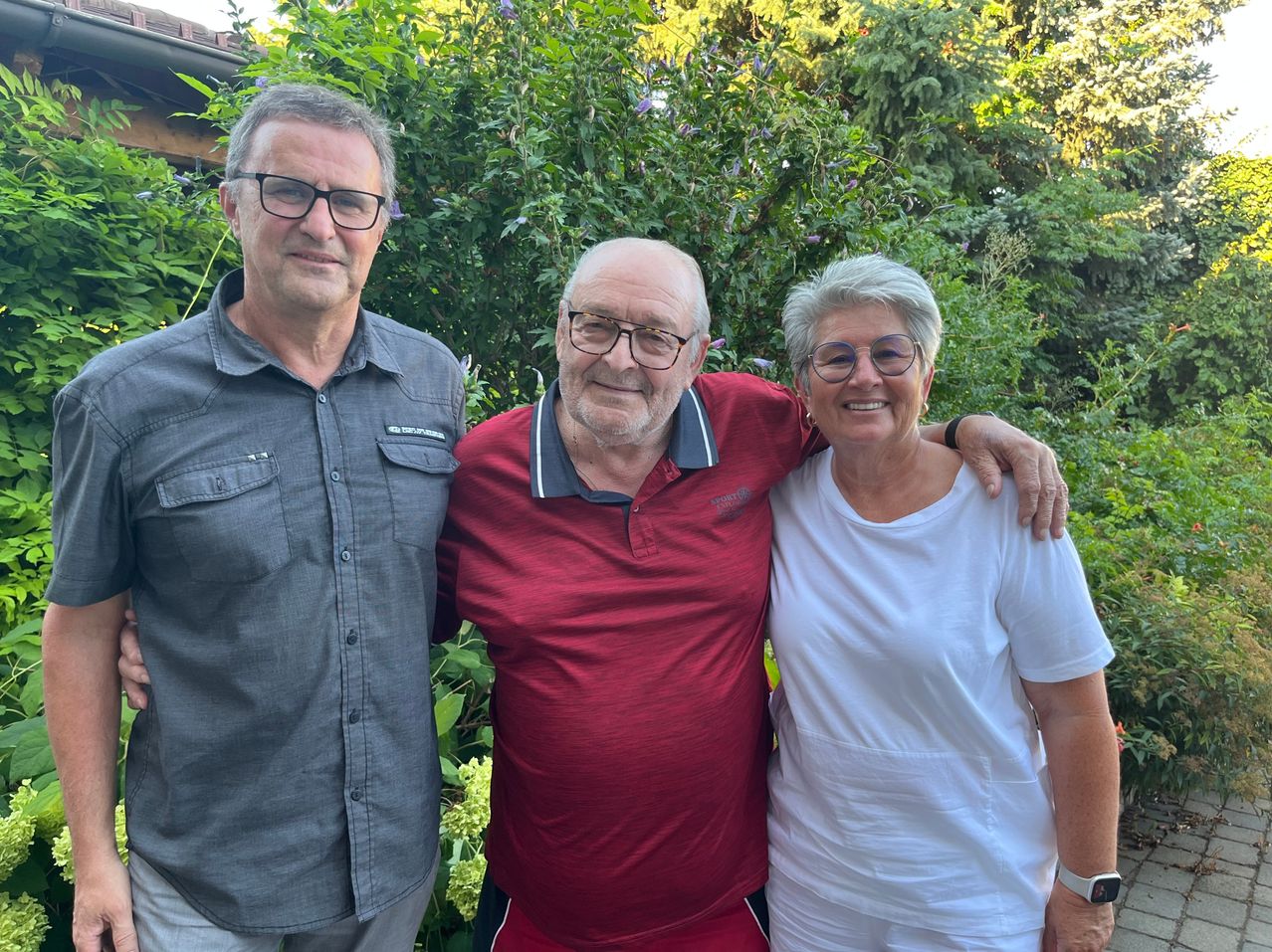 Drei Personen stehen vor grüner Bepflanzung, ein Mann trägt eine graue Hemdbluse, ein älterer Mann eine rote Poloshirt und eine Frau ein weißes T-Shirt.