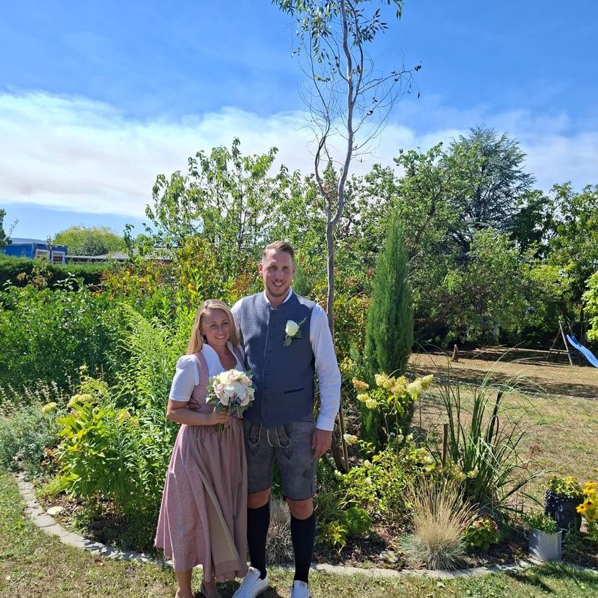 Brautpaar im Garten: Frau im Dirndl mit Blumenstrauß, Mann in Tracht mit Weste und Lederhose.