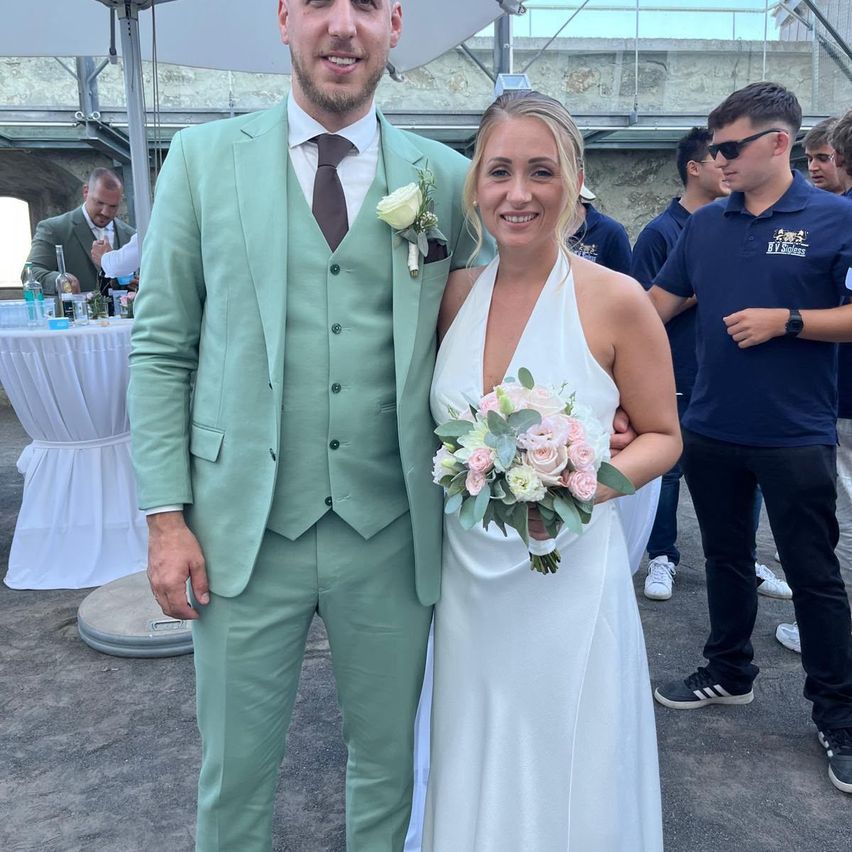 Ein lächelndes Paar steht eng beieinander, der Bräutigam in einem mintgrünen Anzug und die Braut in einem weißen Kleid, mit einem Blumenstrauß. Sie sind von anderen Leuten umgeben, möglicherweise bei einer Hochzeitsfeier.