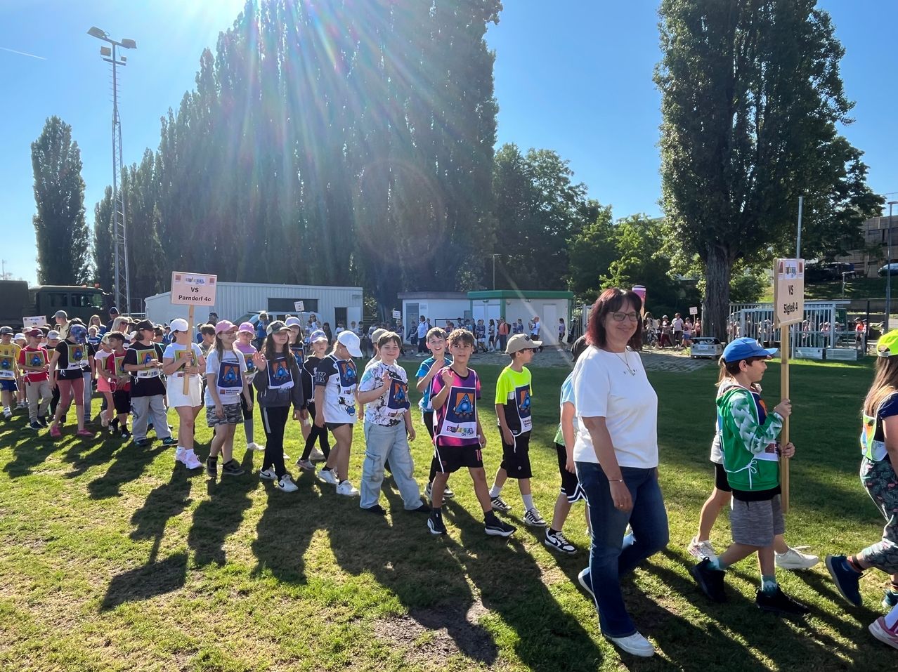 A group of children with colorful shirts are walking on grass in a park, led by a woman with glasses. Behind them, there are trees, buildings, and a crowd of people.