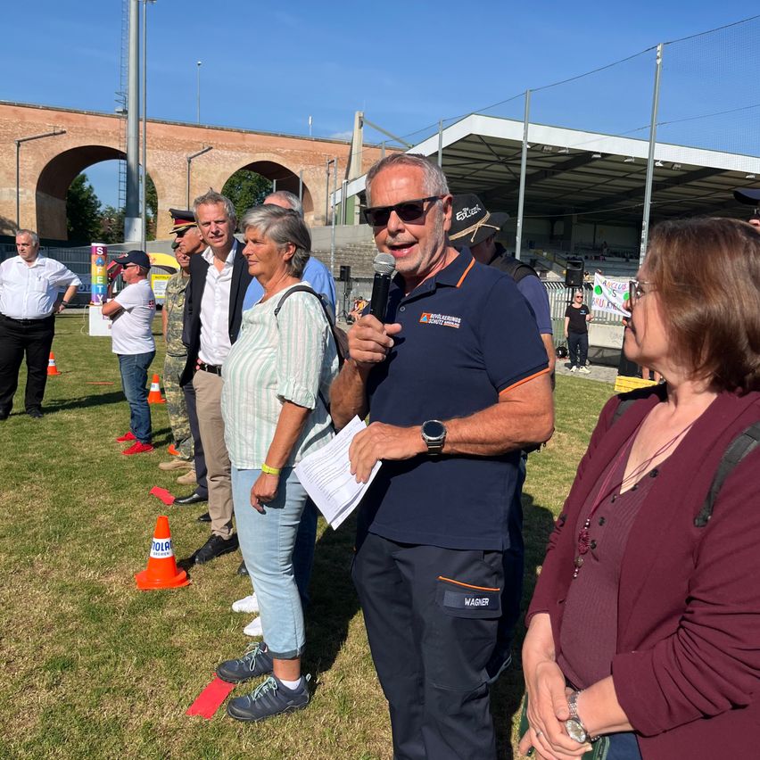 A man in a blue polo shirt stands on a field with a microphone, speaking to a group of people, including a woman in a maroon blazer. Behind them are various orange traffic cones.