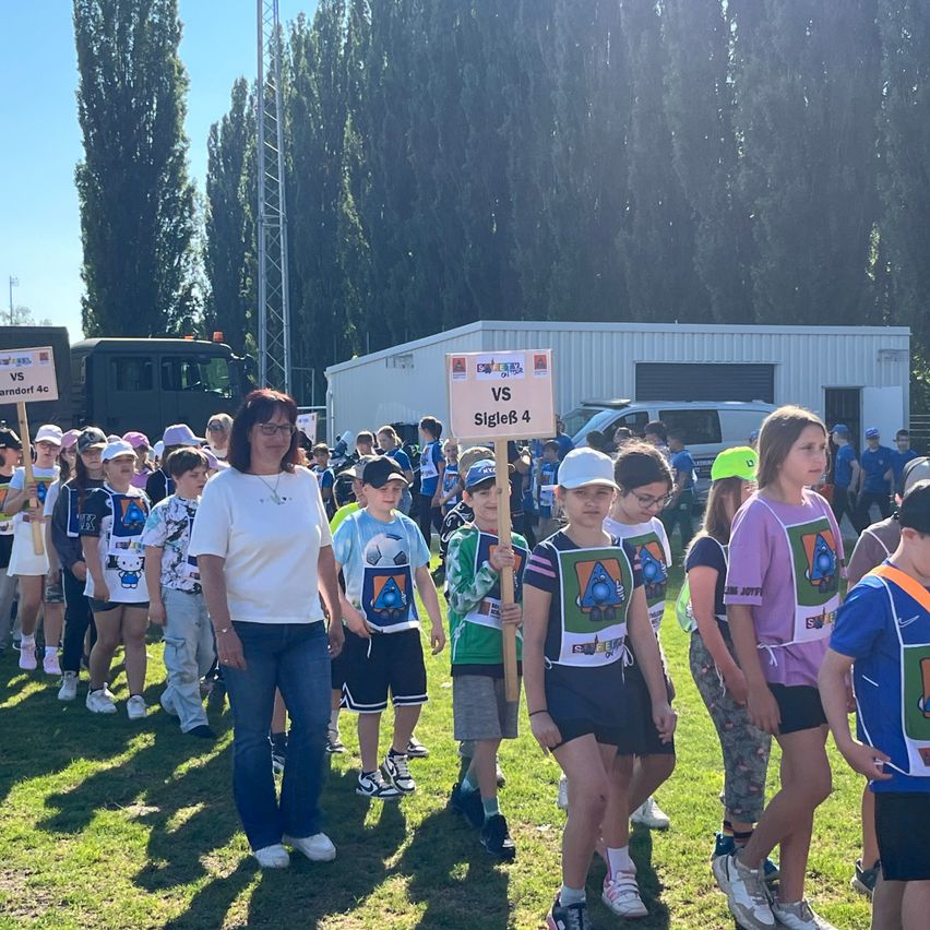 A group of children in matching shirts holding signs with team names walk past adults on a grassy field, with a truck and building in the background.